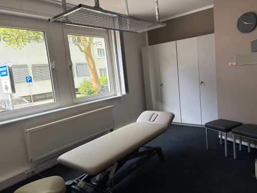 Behandlungsraum mit Massagetisch, Stühlen und großen Fenstern.