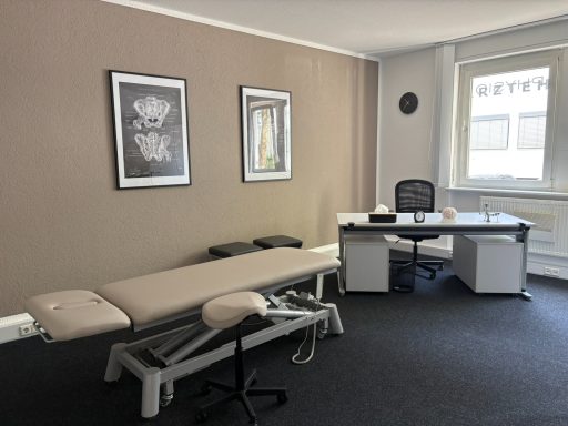 Behandlungsraum mit einer Liege, Schreibtisch und zwei Rahmen mit Kunstwerken an der Wand.